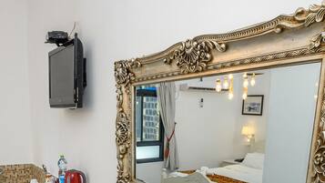A hotel room with a large ornate mirror, a flat-screen TV mounted on the wall, a bathroom with a tiled shower area, and a bed with white bedding.