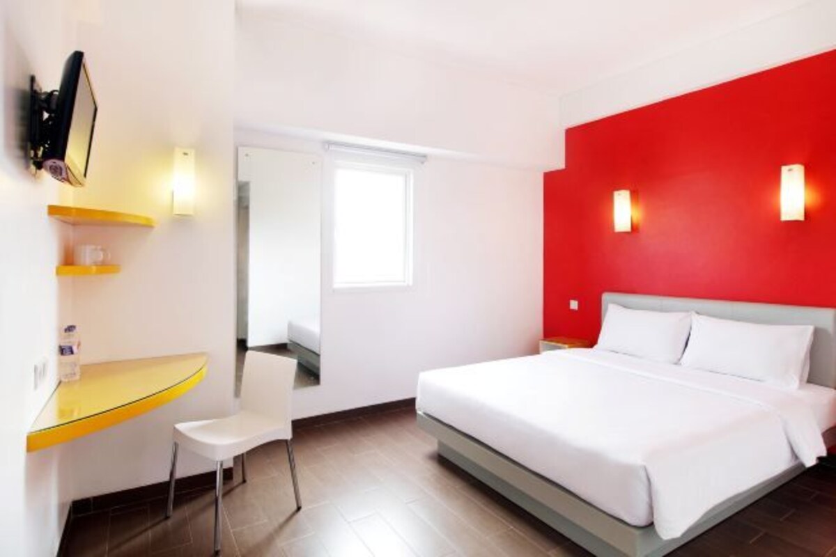 A hotel room with a bed, a desk with a chair, a TV, and a red accent wall.