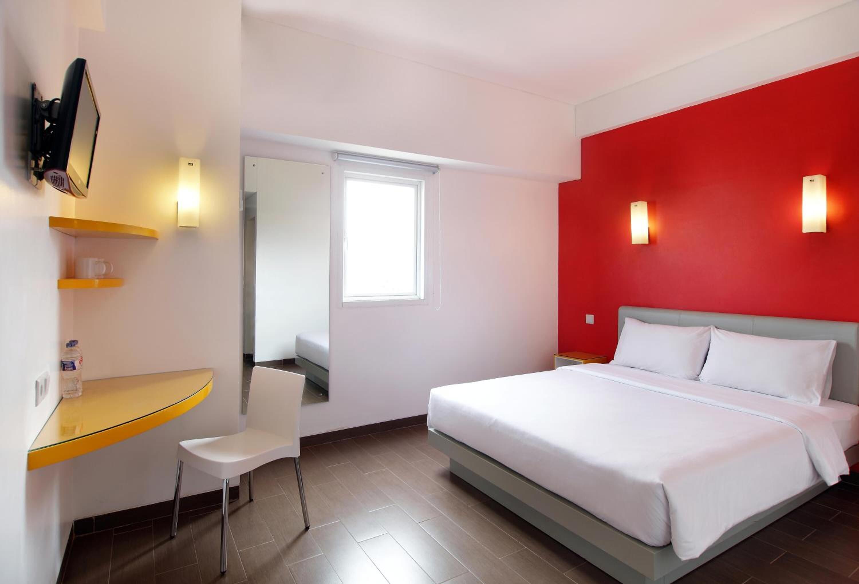 A hotel room with a bed, a desk with a chair, a TV, and a red accent wall.