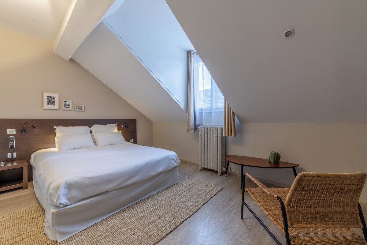 A modern hotel room with a large bed, a small table, a chair, a radiator, and a skylight.