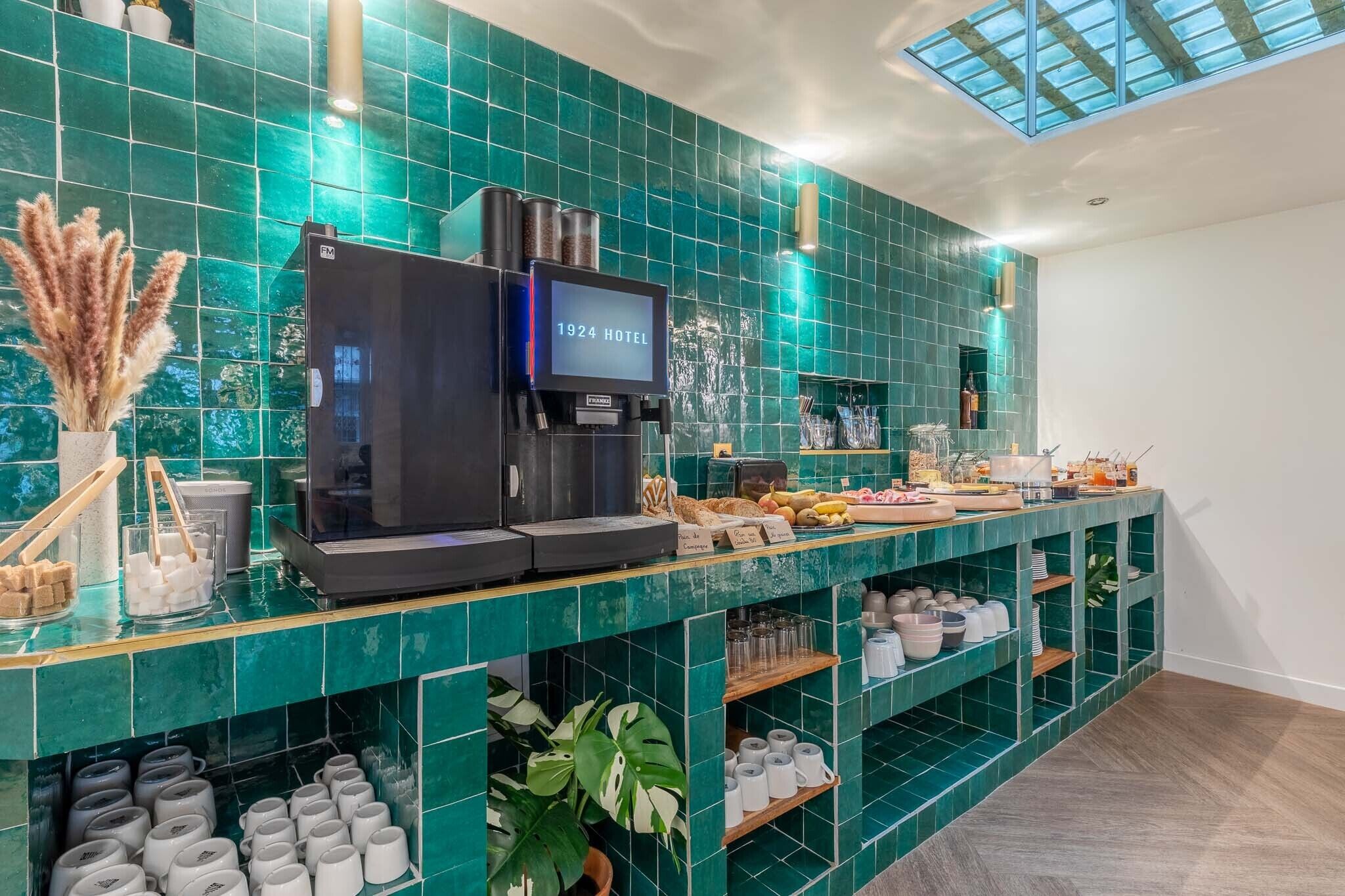 Zona de bar de hotel con un revoque de azulejos verdes, una máquina de café y una variedad de alimentos y bebidas en la barra.