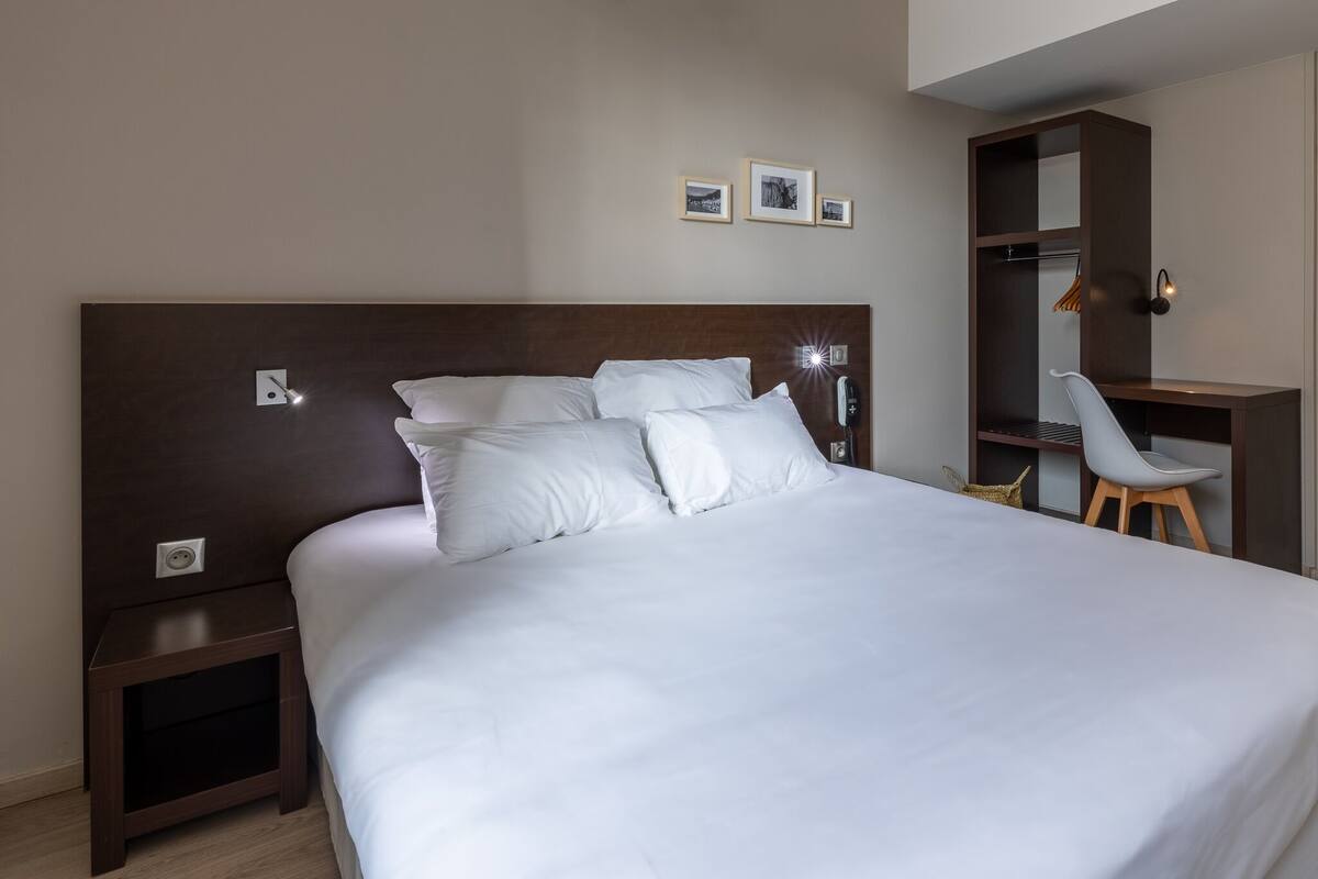 A neatly made bed with white linens, a wooden headboard, a bedside table, and a small shelf with a lamp.