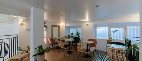 A spacious room with a wooden floor, a dining area with round tables and chairs, and a balcony with plants.