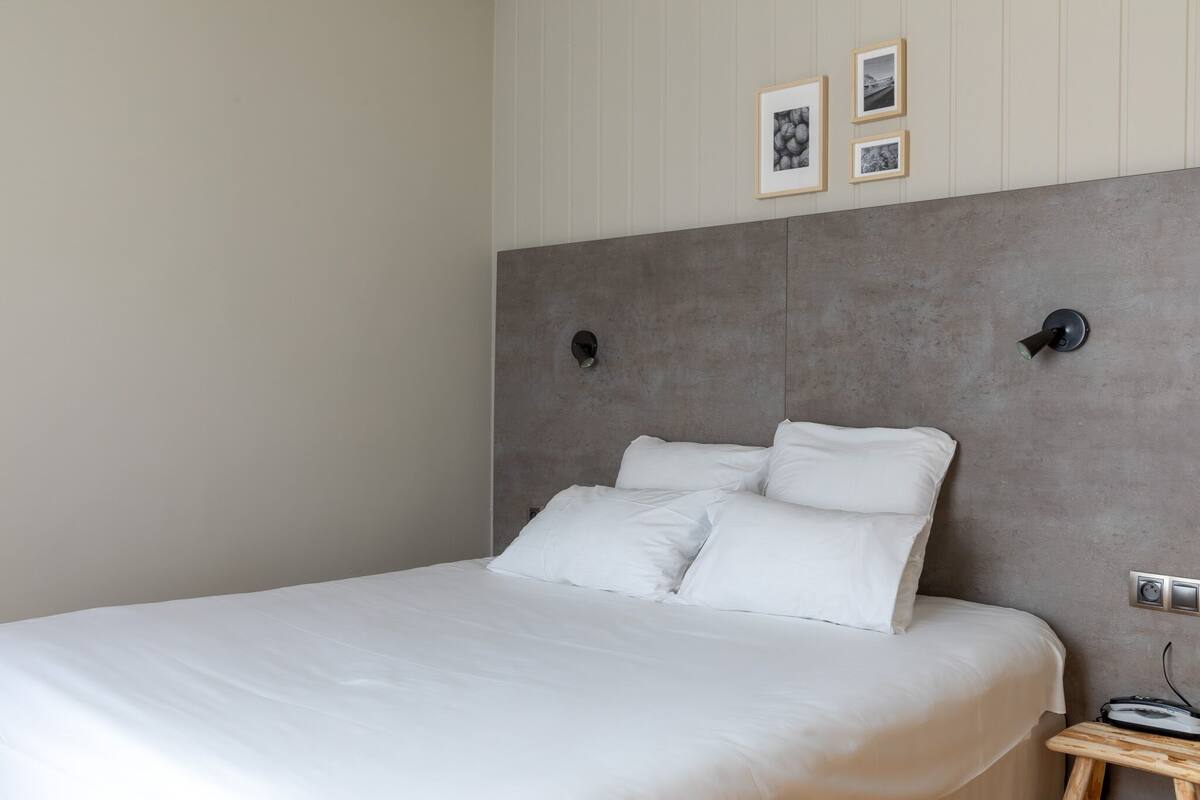 A neatly made bed with white linens in a modern room with a grey headboard, a small bedside table, and framed pictures on the wall.