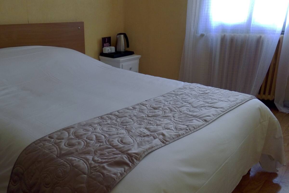 Una cama bien hecha con un edredón blanco y marrón, un cabecero de madera, una mesita de noche con una tetera y una ventana con cortinas.
