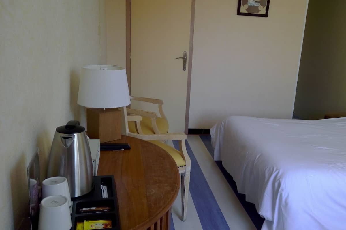 Habitación de hotel con cama, escritorio con lámpara, una silla y una mesita con tazas y un termo.