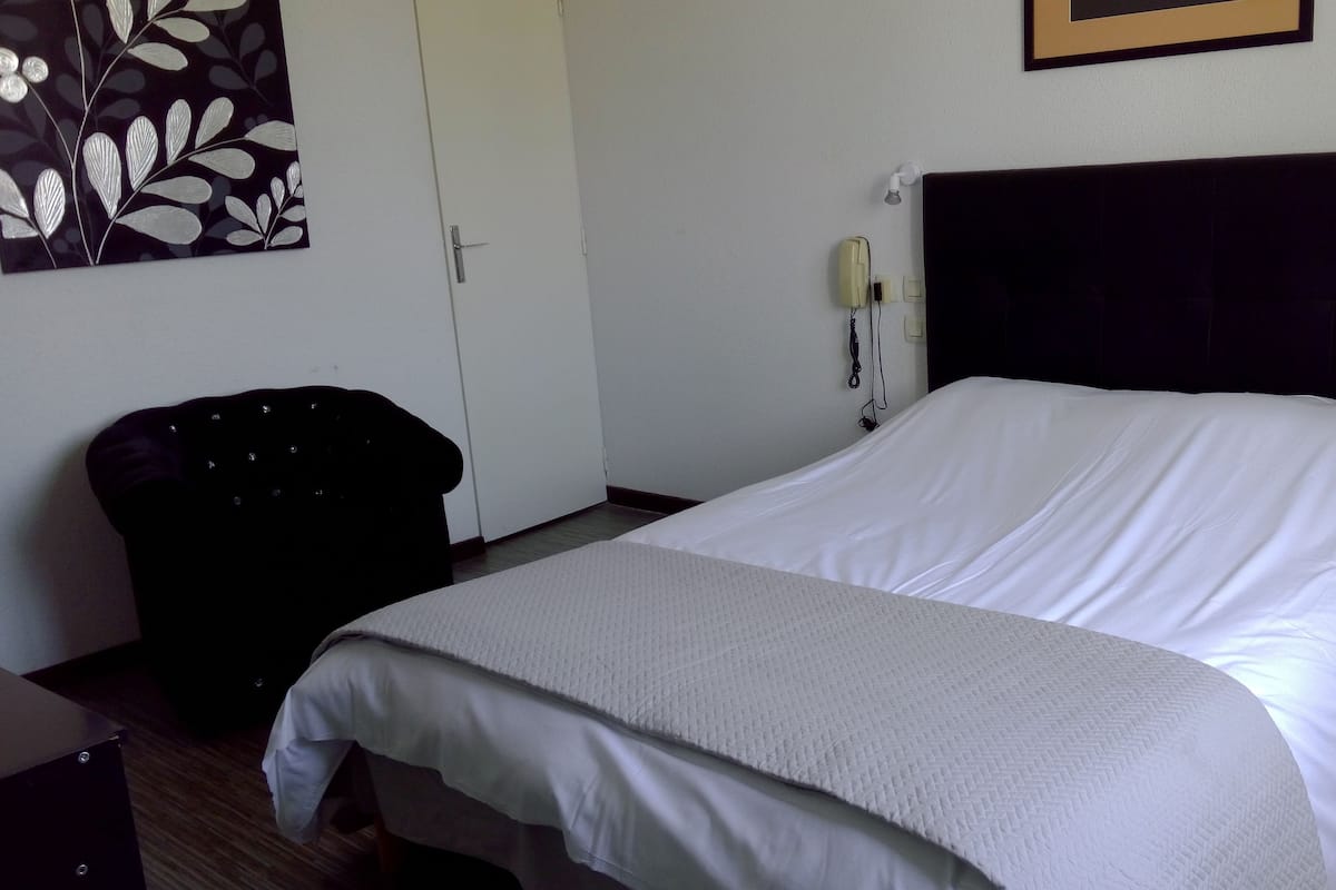 Habitación de hotel con cama, una silla, una mesita de noche, un teléfono en la pared y un cuadro enmarcado.