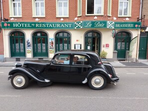 Ein klassisches, schwarzes Auto vor einem Gebäude mit Schildern, auf denen "HOTEL RESTAURANT", "La Paix" und "BRASSERIE" stehen.
