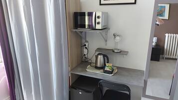 A compact room with a desk, a kettle, a chair, a shelf with a lamp, and a radiator.
