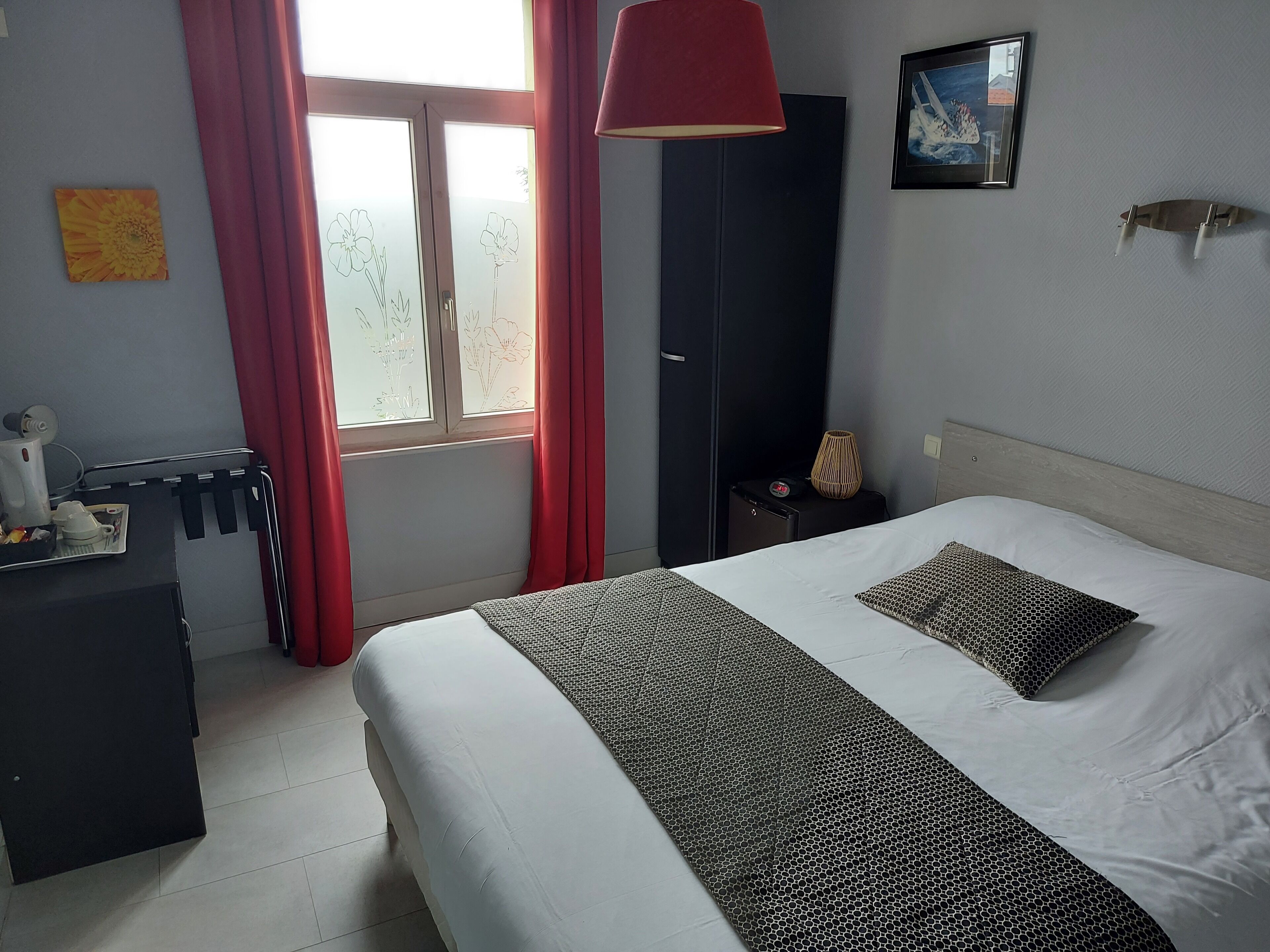 Habitación de hotel con cama, mesita de noche, escritorio, silla, ventana con cortinas y lámpara de pared.