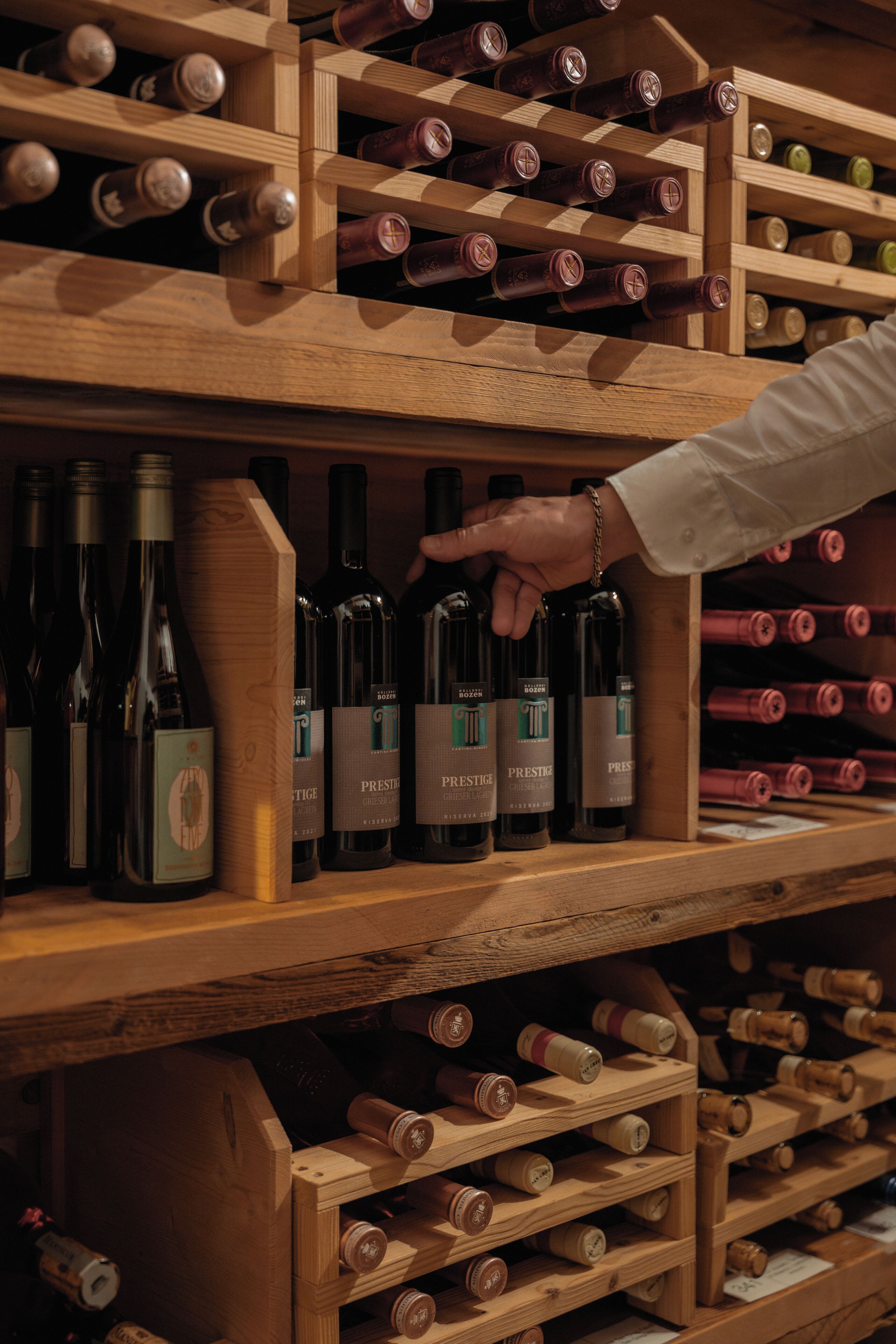 Ein Weinschränk aus Holz mit Flaschen, eine Hand, die nach einer Flasche greift, und Etiketten mit dem Wort 'PRESTIGE'.