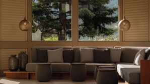 Een moderne woonkamer met een bank, poefjes en decoratieve vazen, grote ramen met uitzicht op bomen, en hanglampen.