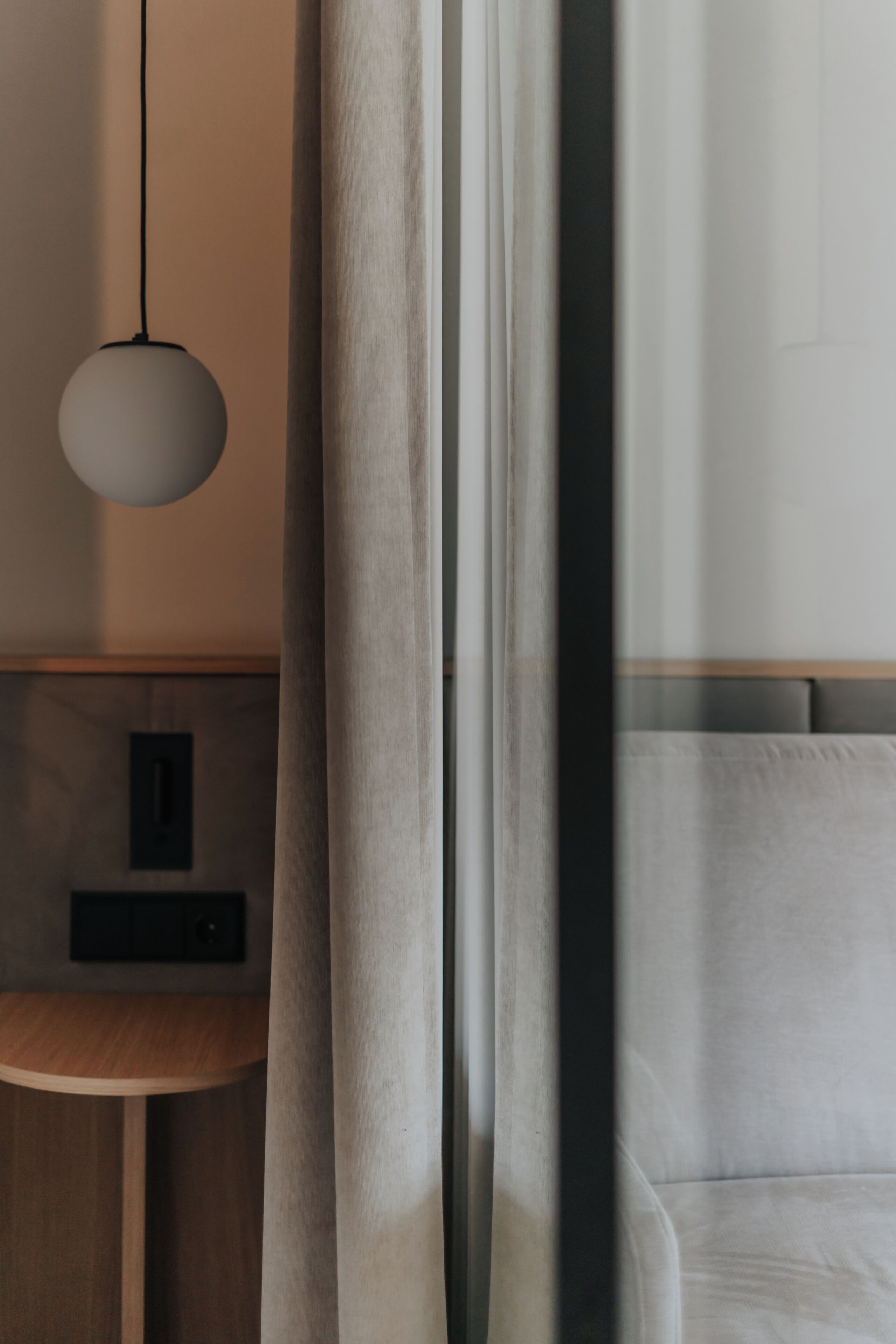 A modern room with a bed, a round pendant light, a small wooden table, and a curtain partially drawn.