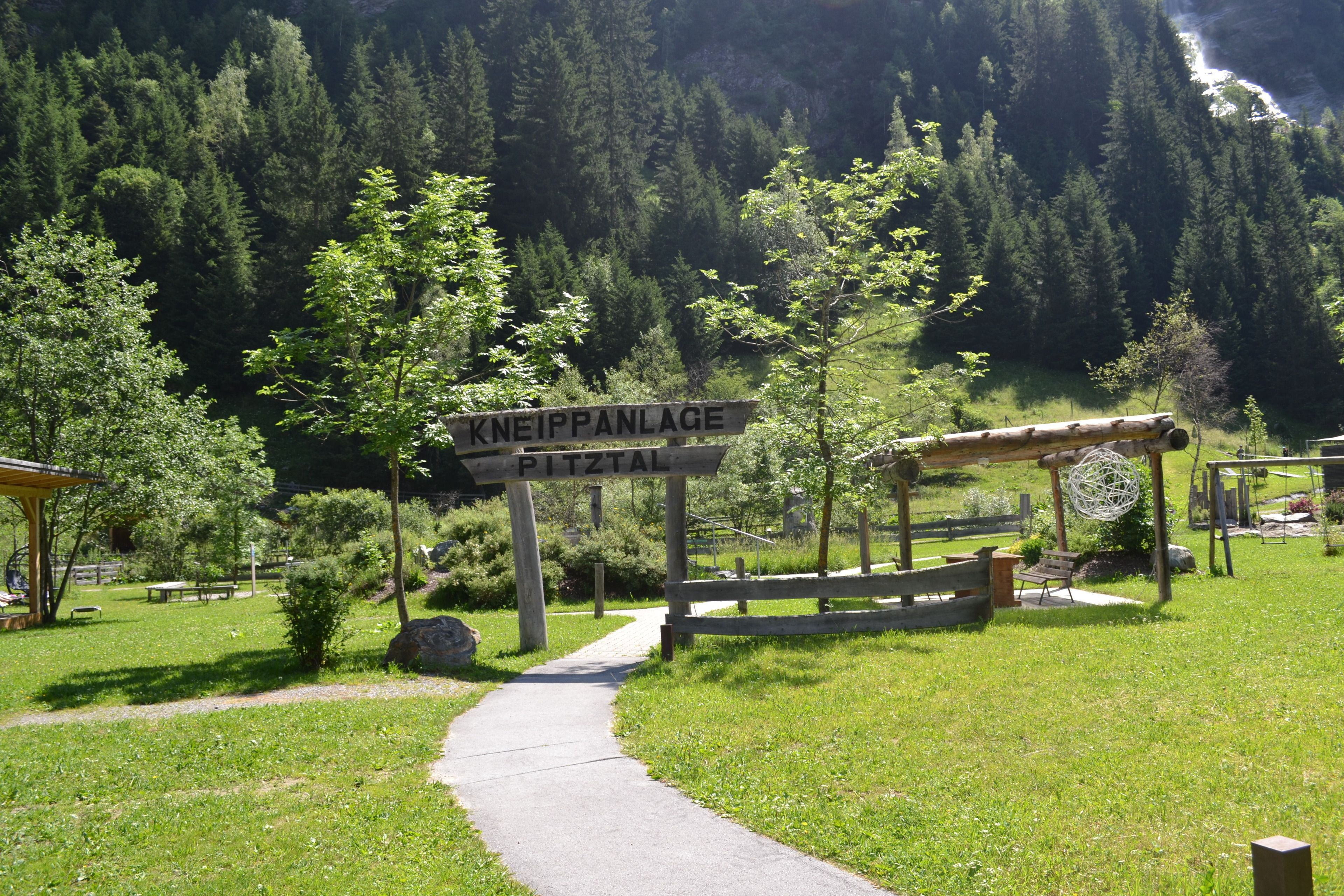 A pathway leading to a signpost with 'Kneippanlage' and 'Pitzthal' written on it, surrounded by greenery and a mountainous backdrop.