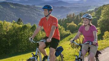 Duas pessoas de bicicleta numa ciclovia, com vista panorâmica de colinas e árvores.