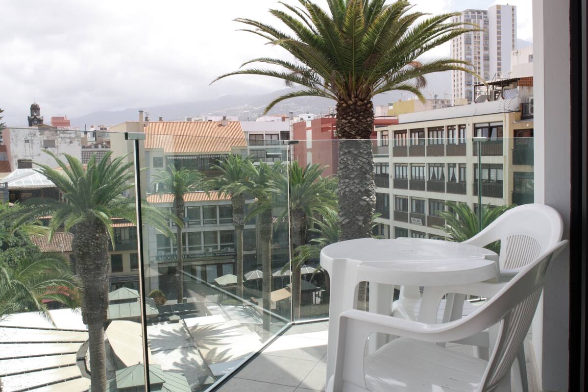 A balcony with a glass railing, white plastic chairs, and a palm tree.