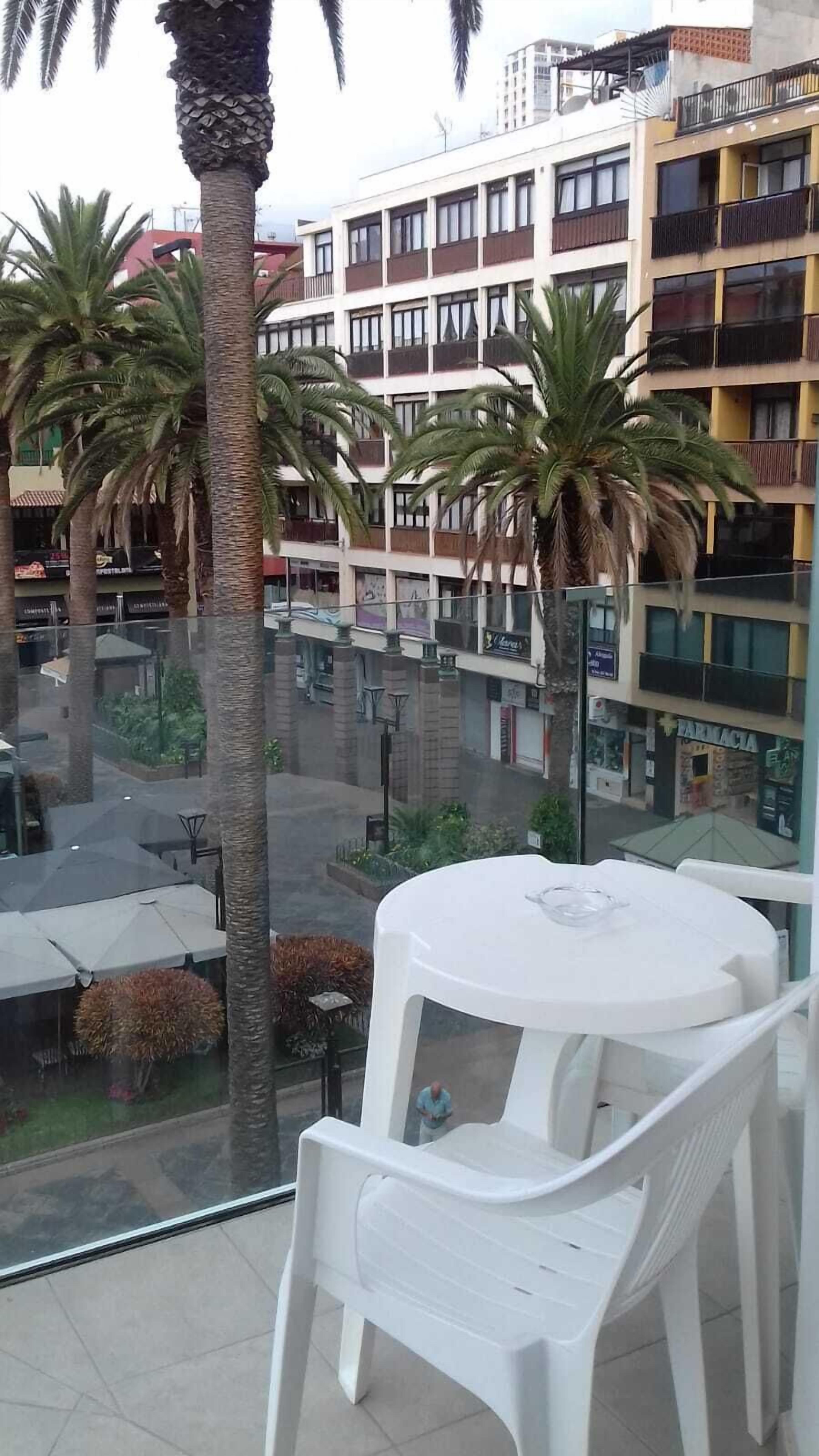 Un balcón con sillas y una mesa de plástico blanco, con vista a una calle con palmeras y un edificio de varios pisos con balcones.