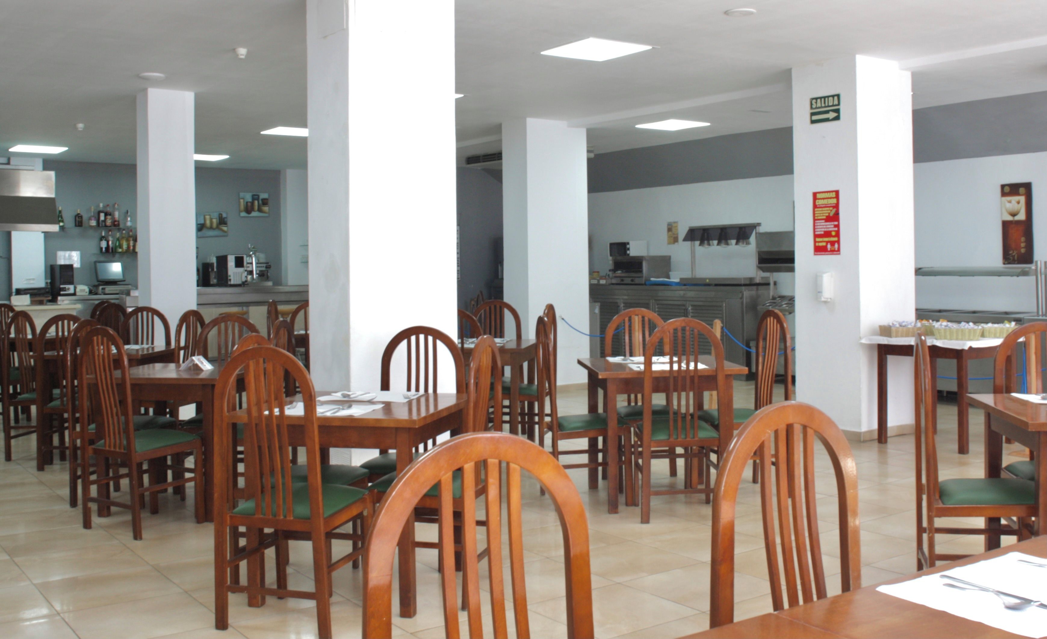 Un comedor amplio con mesas y sillas de madera, una cocina al fondo y un letrero que indica la salida.