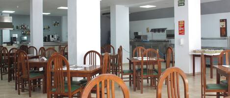 Un comedor amplio con mesas y sillas de madera, una cocina al fondo y un letrero que indica la salida.