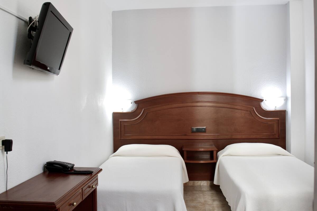 A hotel room with a wooden headboard, two beds, a bedside table with a phone, and a flat-screen TV mounted on the wall.
