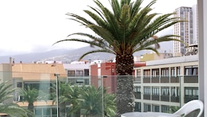 A balcony with a glass railing, a white plastic table and chairs, a palm tree, and a view of buildings and a mountain in the background. - Tropical (Puerto de la Cruz)
