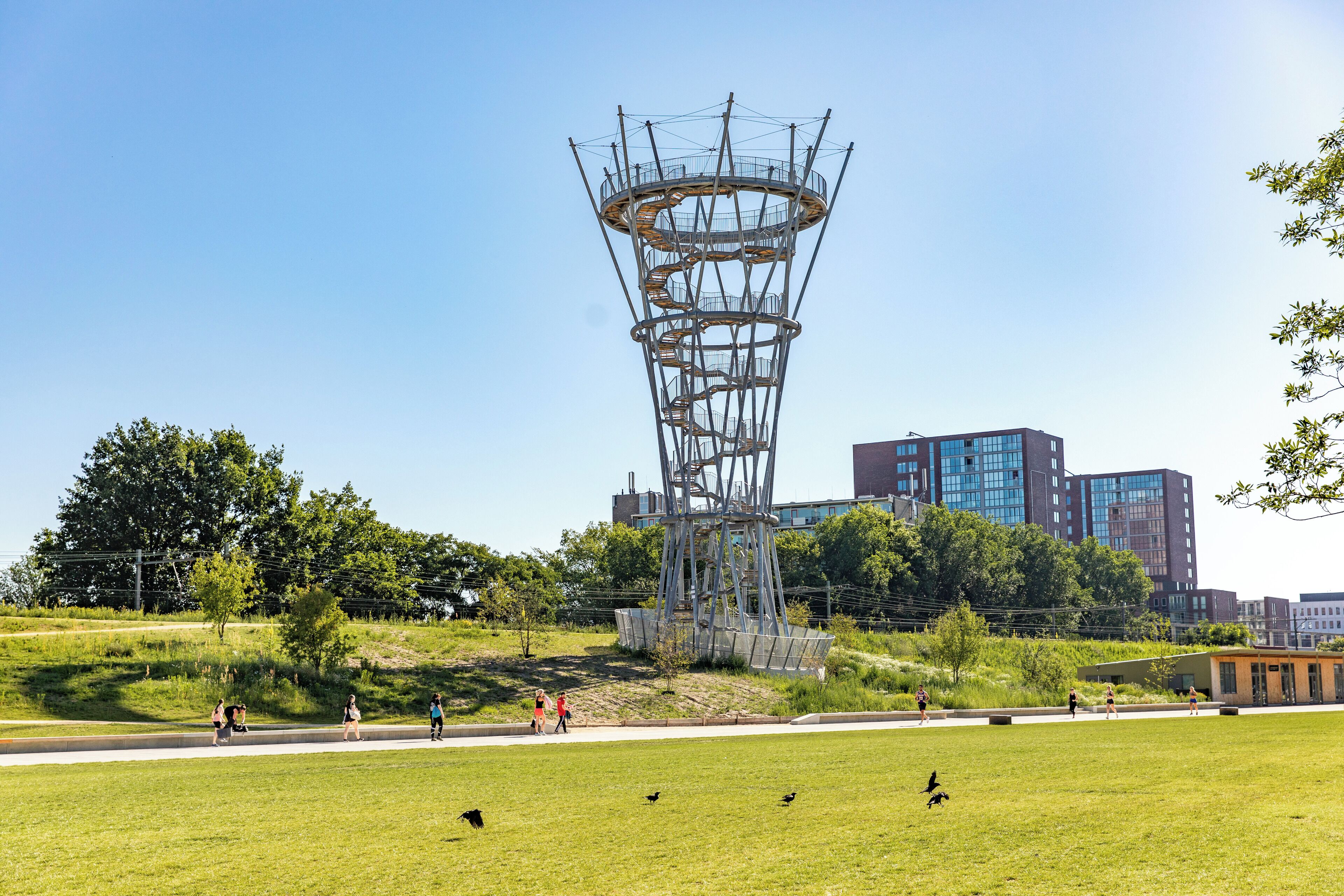 A modern tower structure in a park with a clear sky, surrounded by greenery and a few people walking.