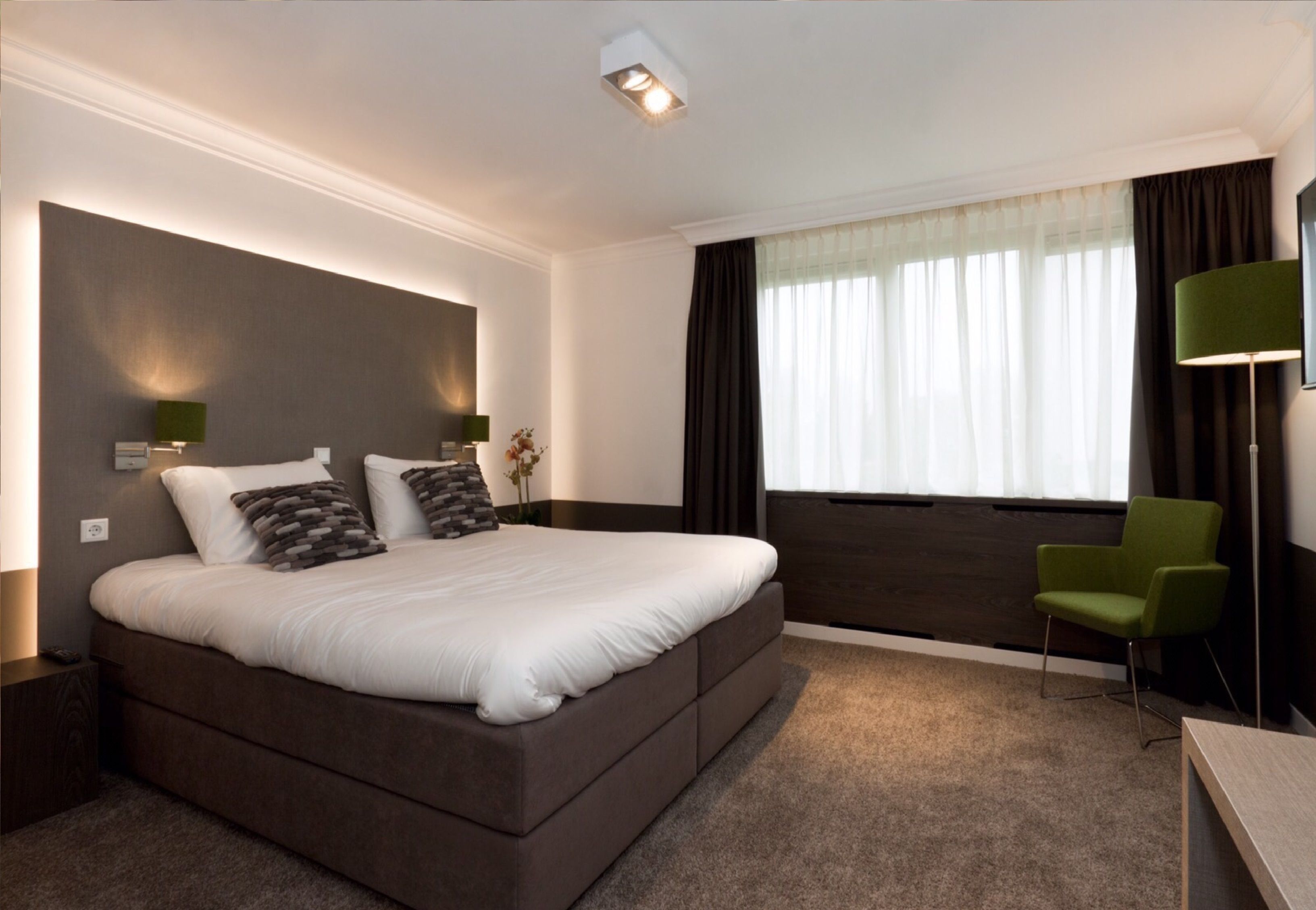Una habitación de hotel moderna con una cama grande, un sillón verde, un escritorio y una ventana con cortinas.
