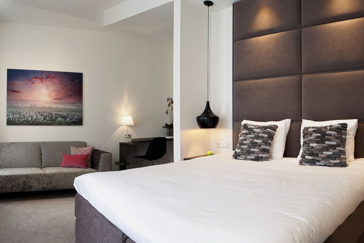 Une chambre d’hôtel moderne dotée d’un grand lit, d’un canapé, d’un téléviseur à écran plat et d’un tableau représentant un coucher de soleil.