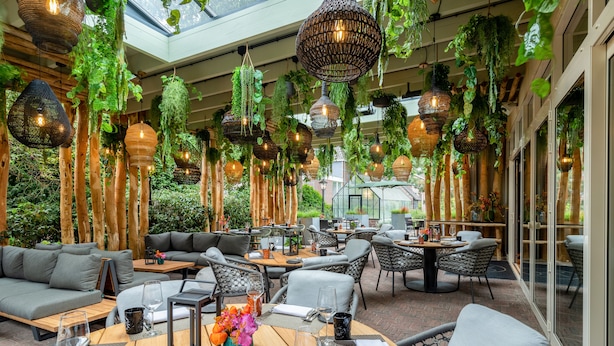 Un comedor con espacio interior y exterior, con lámparas colgantes de mimbre, asientos acolchados y una mesa de madera con un jarrón de flores.