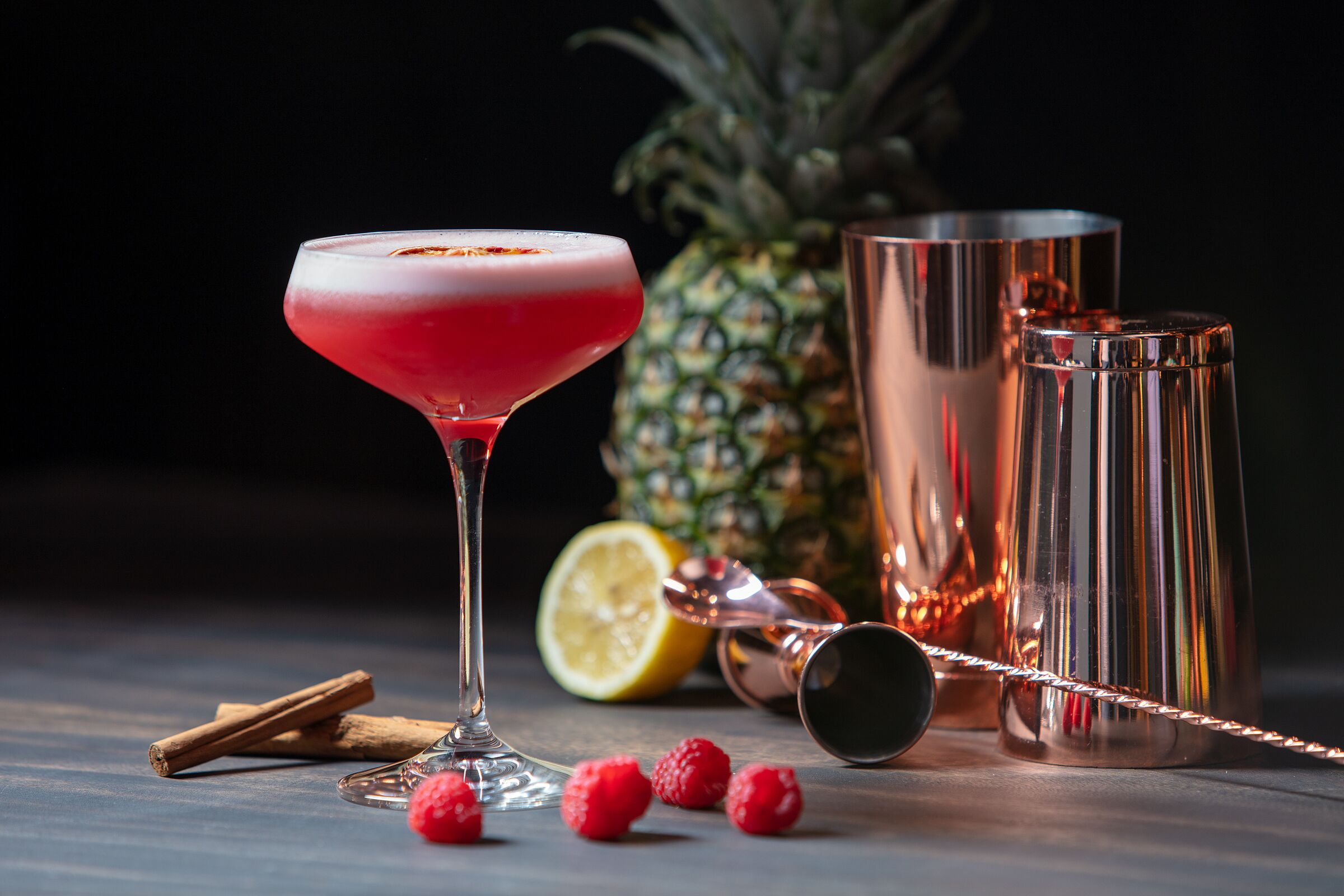 Un vaso de cóctel con una bebida roja, una piña, media lima y algunas frambuesas sobre una mesa.