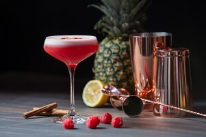 A cocktail glass with a red drink, a pineapple, a lemon half, and some raspberries on a table.