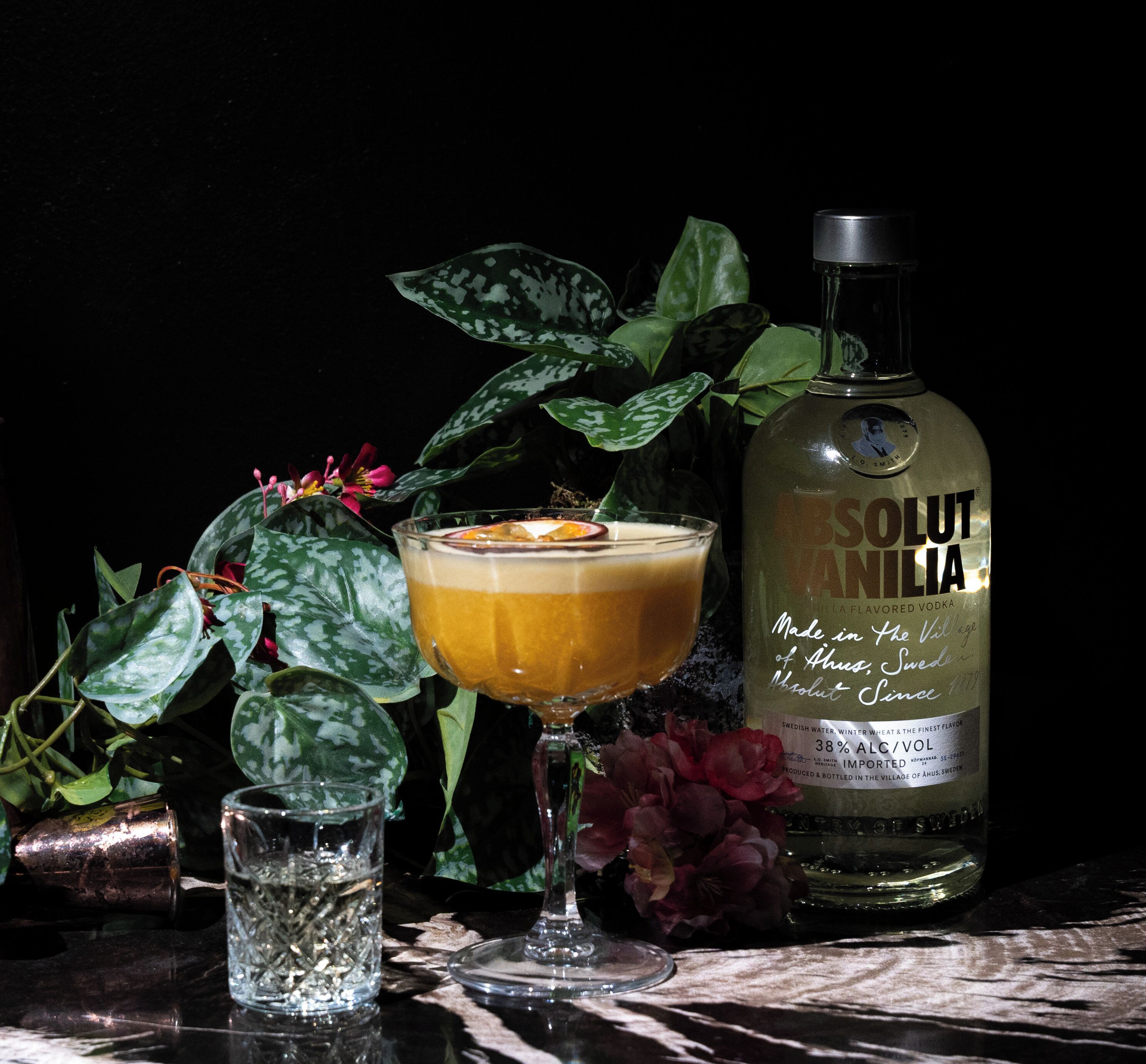 A bottle of Absolut Vanilla vodka, a glass of cocktail with an orange slice garnish, and a small glass of water on a dark surface with green plants and flowers.