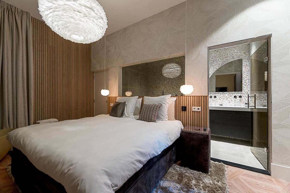 Une chambre moderne avec un grand lit, une armoire intégrée, un miroir et une salle de bain visible à travers une porte en verre.