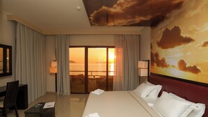 Ein Hotelzimmer mit einem großen Bett, einem Schreibtisch, einem Sessel und Blick auf den Sonnenuntergang durch die Glastür.