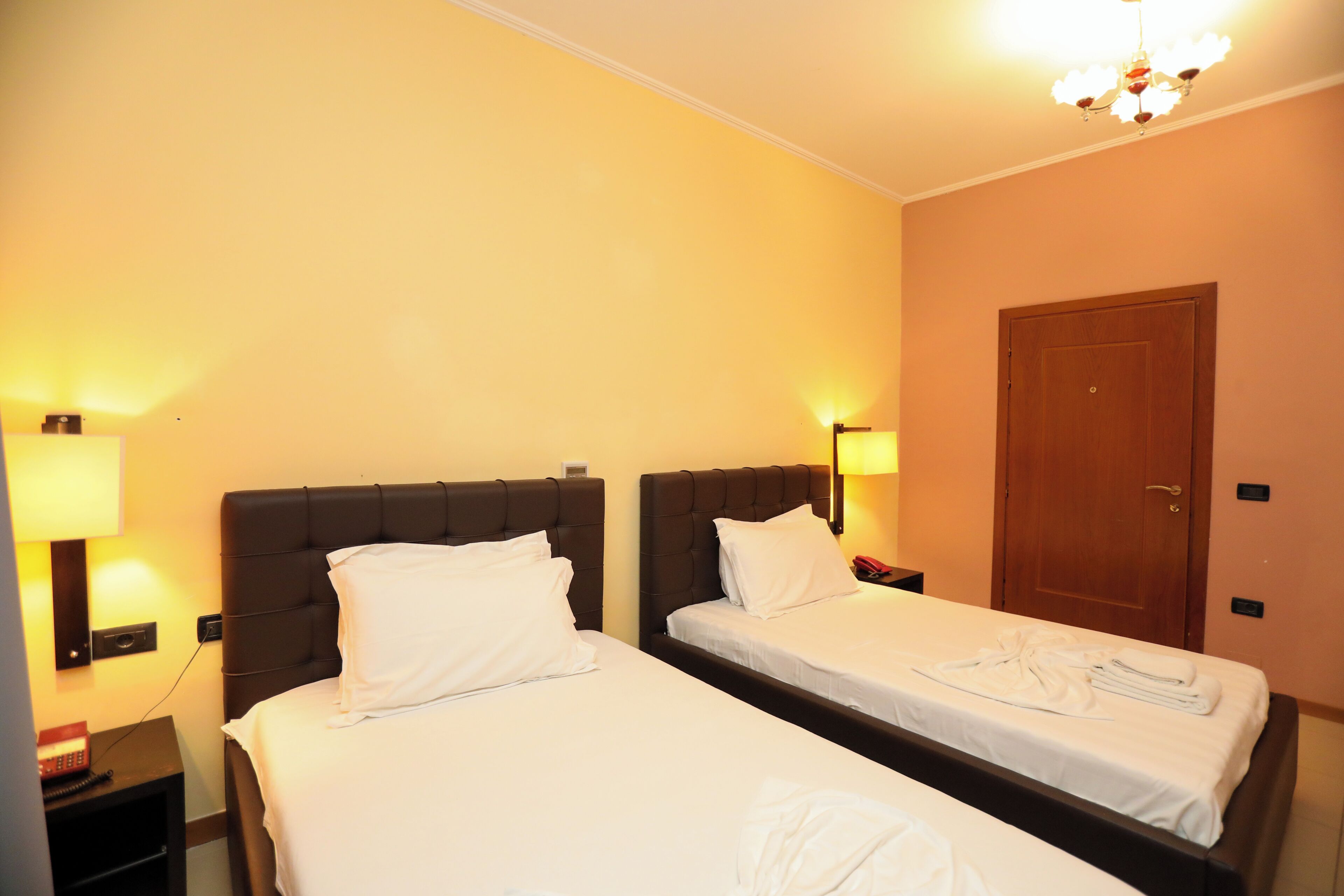 Habitación de hotel con dos camas, cada una con sábanas blancas y cabeceros oscuros. Un puerta de madera y una lámpara de pared a cada lado de las camas.