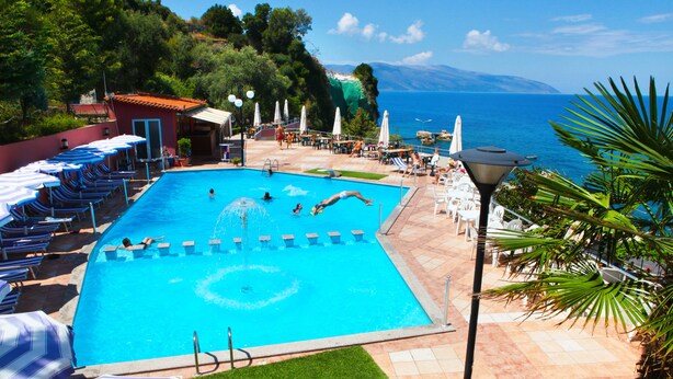 Ein Schwimmbecken mit Liegestühlen und Sonnenschirmen, umgeben von einer angelegten Grünfläche mit Blick auf das Meer.