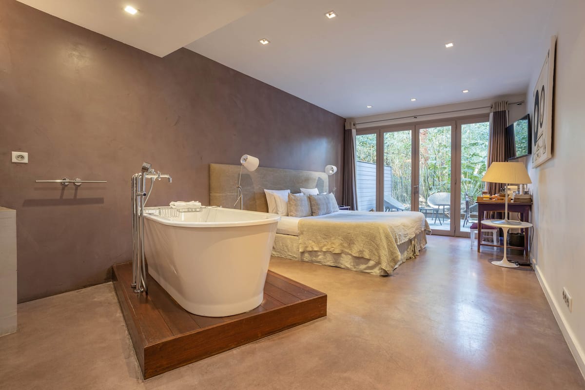 Un dormitorio moderno con una bañera grande, una cama con cabecera y vistas al exterior a través de amplias ventanas.