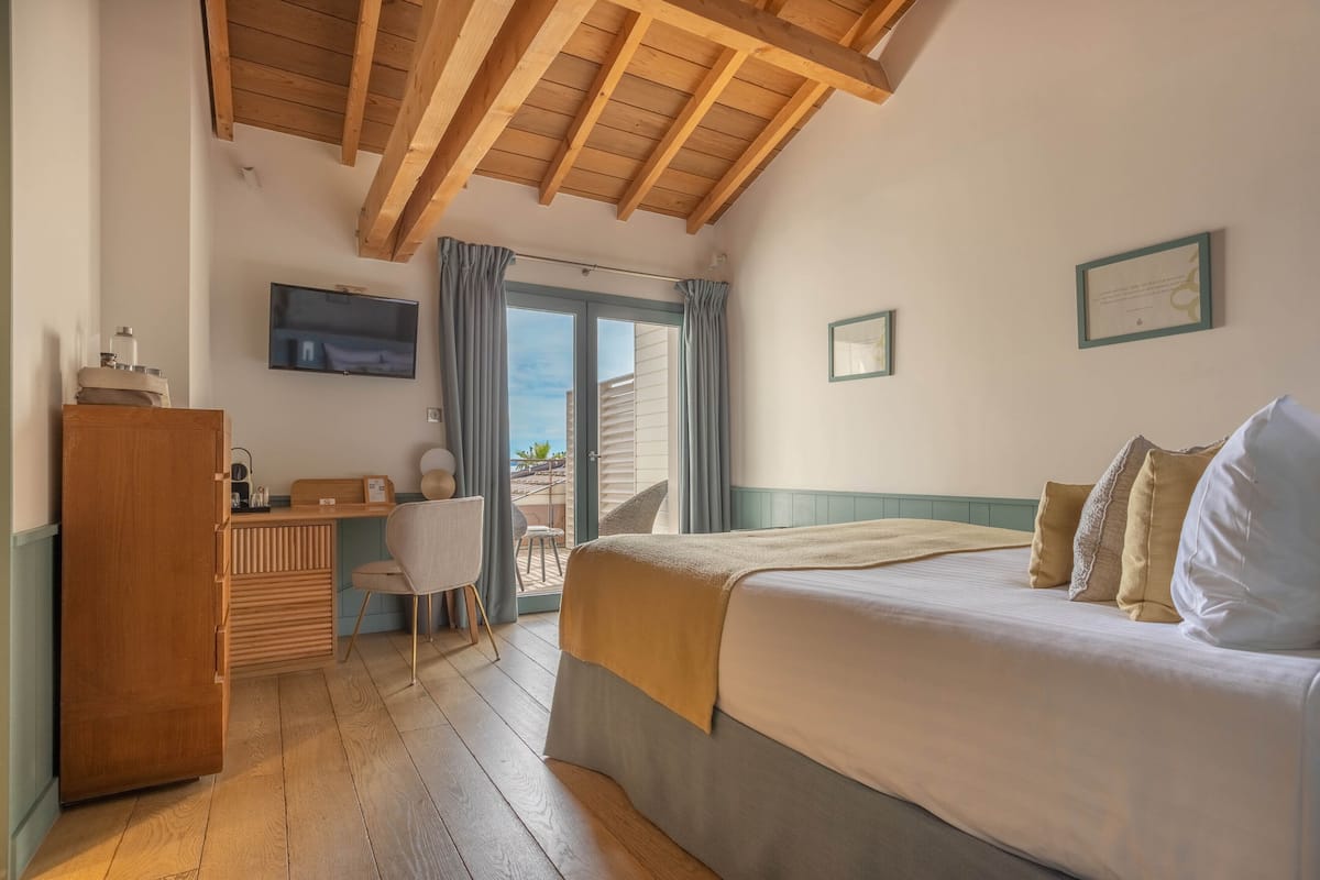 Habitación de hotel con cama, escritorio, silla, televisor y balcón con cortinas.