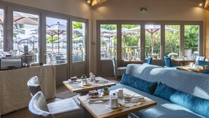 Un restaurante con una decoración moderna, que presenta un sofá azul, mesas de madera y grandes ventanales con vistas a una terraza.