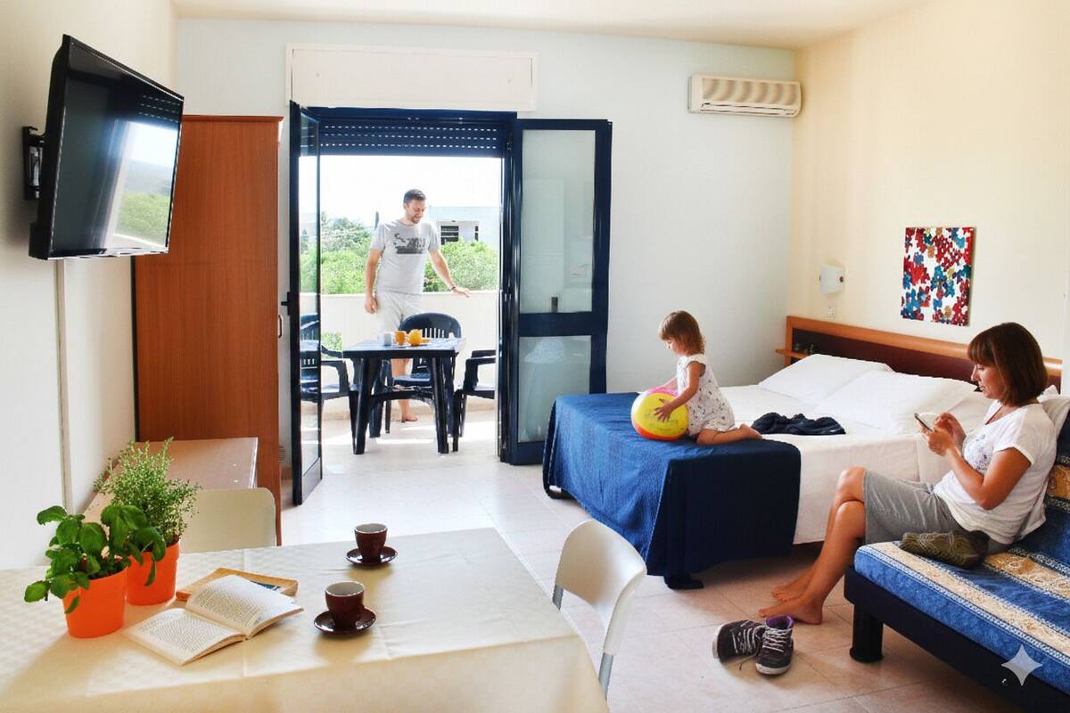 Ein Hotelzimmer mit einem Bett, einem Tisch mit Buch und Tasse, einem an der Wand befestigten Fernseher und einem Balkon mit Tisch und Stühlen.
