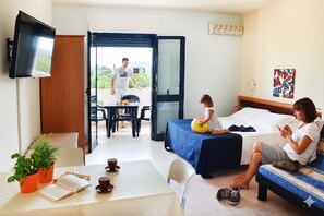 A hotel room with a bed, a table with a book and a cup, a TV mounted on the wall, and a balcony with a table and chairs. - Oasi Salento Residence Hotel (Melendugno)