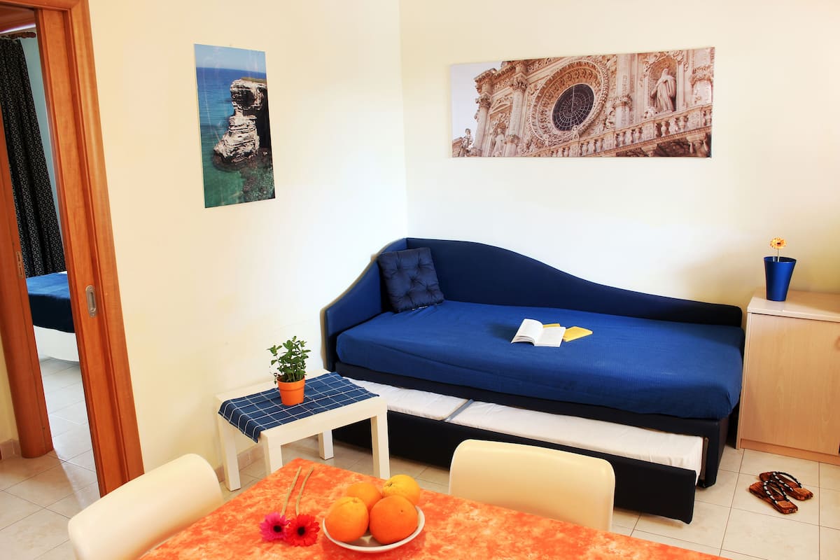 Ein gemütlicher Raum mit einem blauen Sofa, einem kleinen Tisch mit einer Topfpflanze, einer Orangenschale und einem Bett mit blauer Bettdecke.