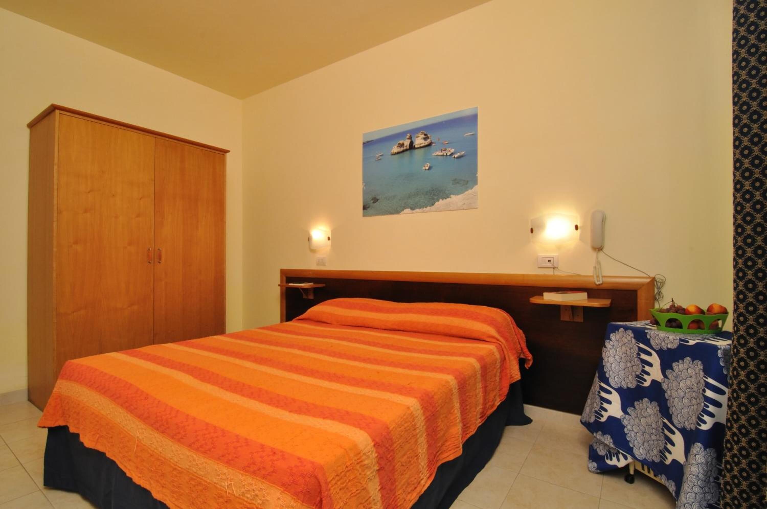 A bedroom with a bed, wooden wardrobe, a desk with a lamp, a painting of a coastal scene, and a table with fruit.