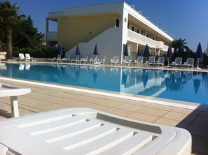 A swimming pool with white lounge chairs and blue umbrellas, surrounded by a building with balconies. - Oasi Salento Residence Hotel (Melendugno)