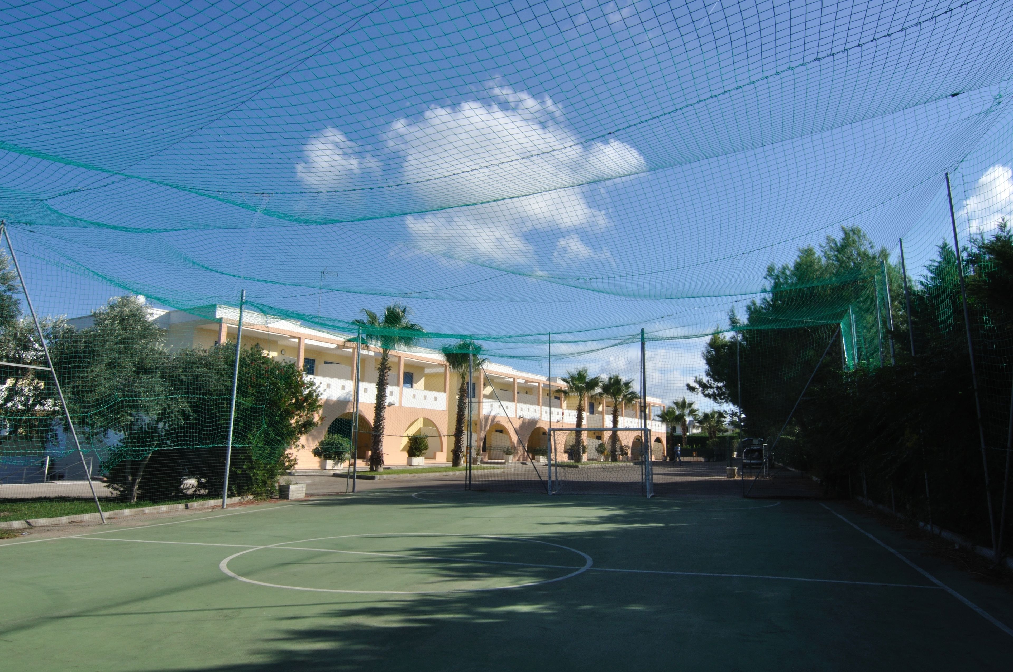 A tennis court with a net and surrounding netting, a building with arches and palm trees in the background.