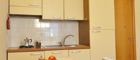 Una pequeña kitchenette con un lavamanos, estufa y armarios. Hay una mesa con mantel azul y una cesta de frutas.