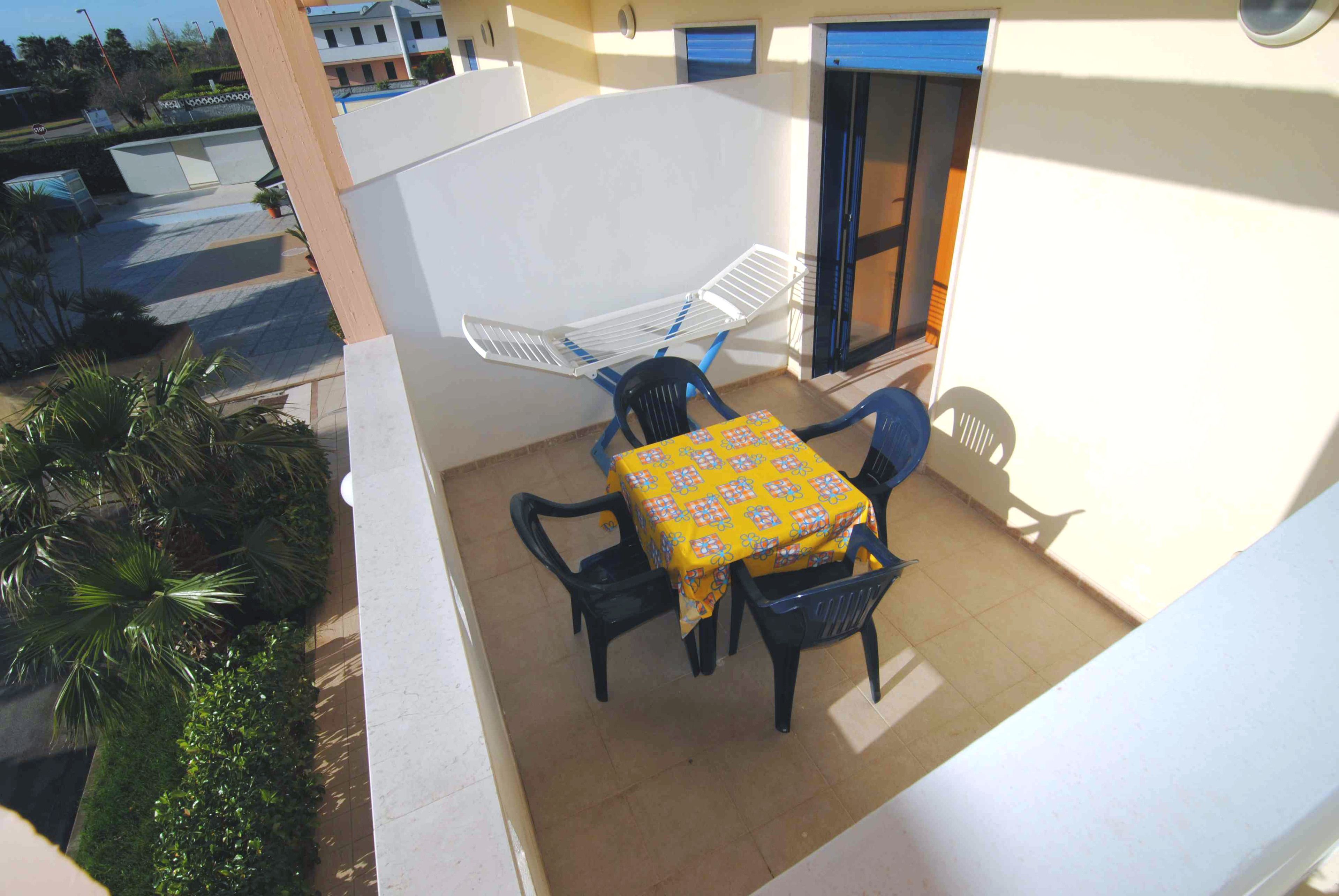 A balcony with a table and chairs, a white chair, and a blue chair.