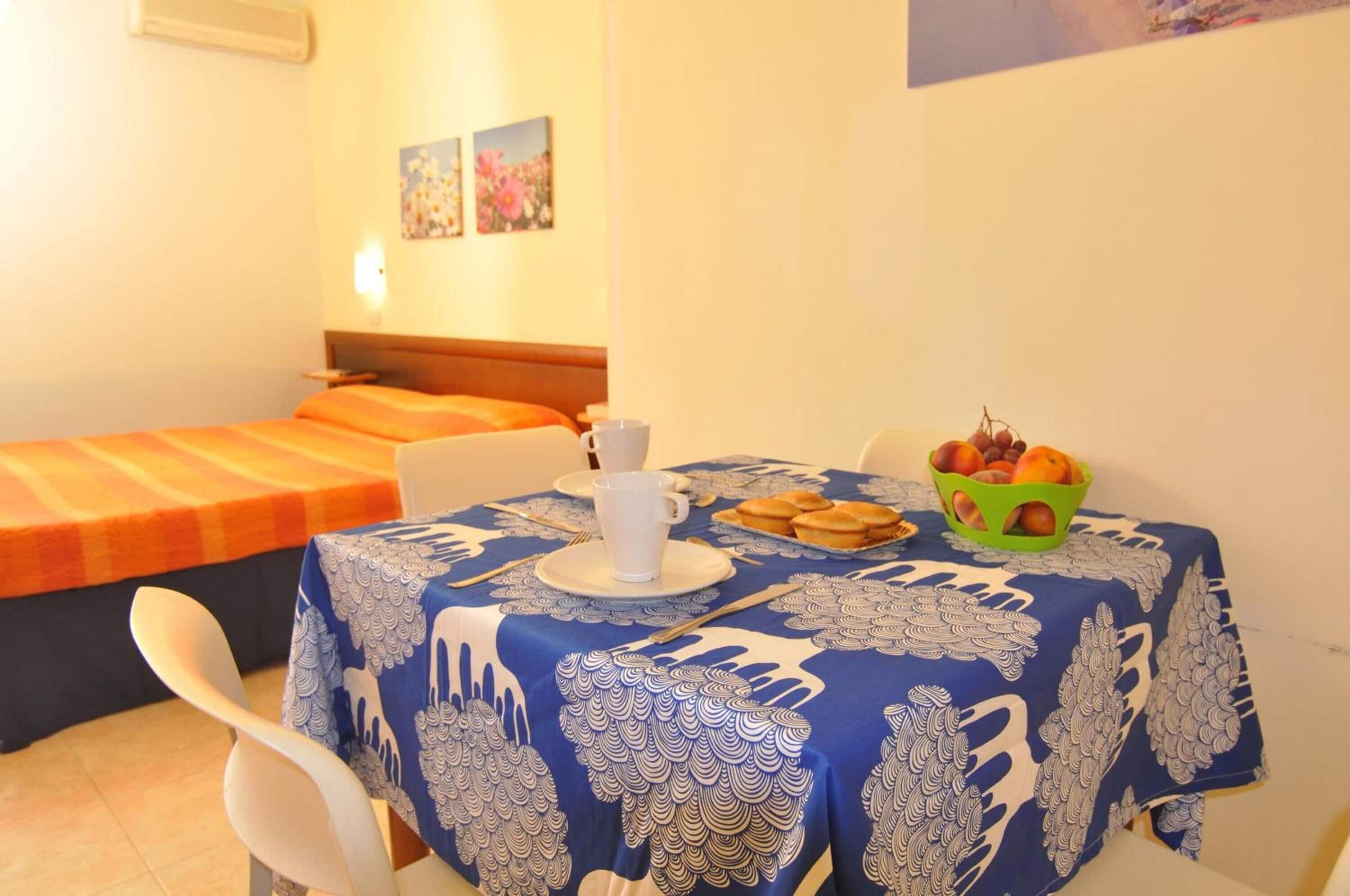 A hotel room with a single bed, a dining table with a blue tablecloth, a bowl of fruit, and a white cup.