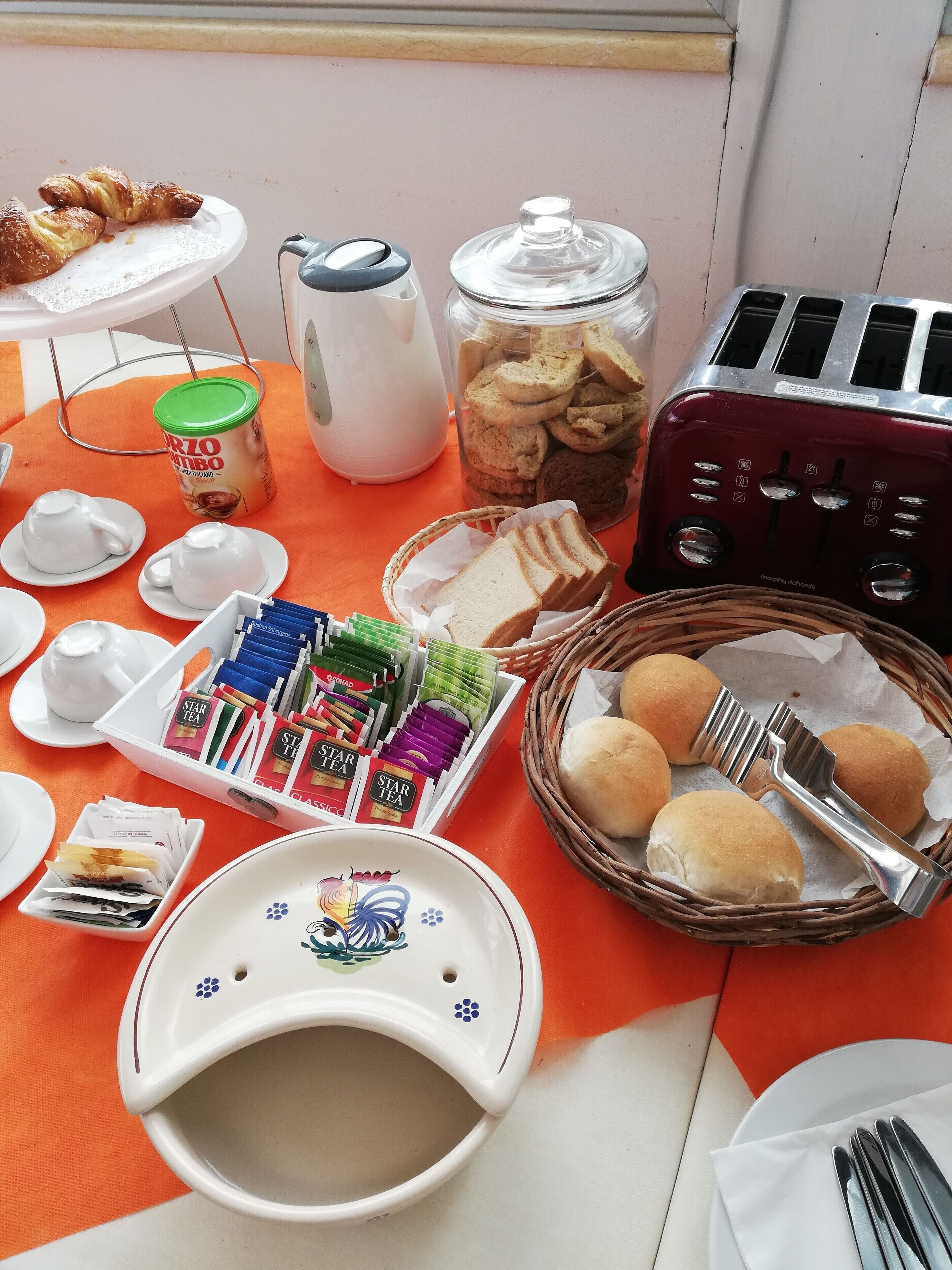 A breakfast setup with croissants, a jar of cookies, a toaster, tea packets, and bread rolls on an orange table.