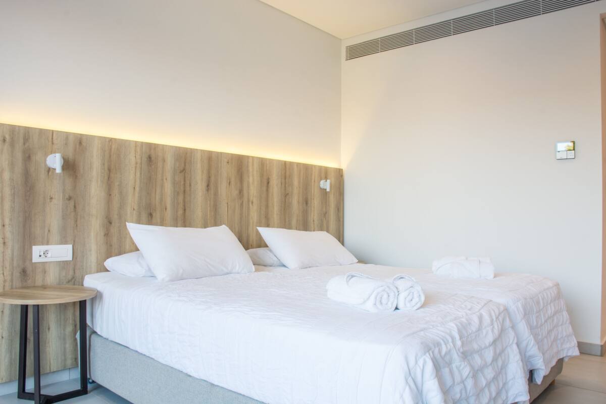 A modern hotel room with a wooden headboard, a round wooden table, and a neatly made bed with white linens and a rolled towel.